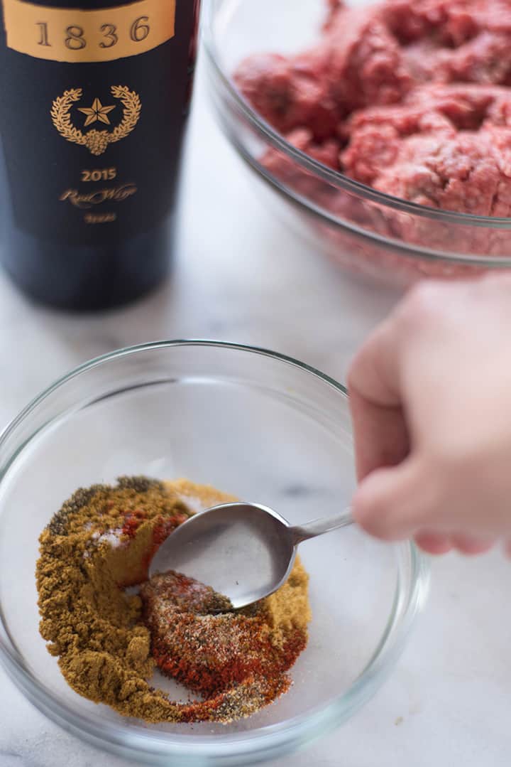 Mixing together the spices for the Texas chili recipe, which include cumin, paprika, sea salt, black pepper, dry mustard, granulated onion, and granulated garlic, alongside a bowl of ground lean beef and a bottle of red wine.