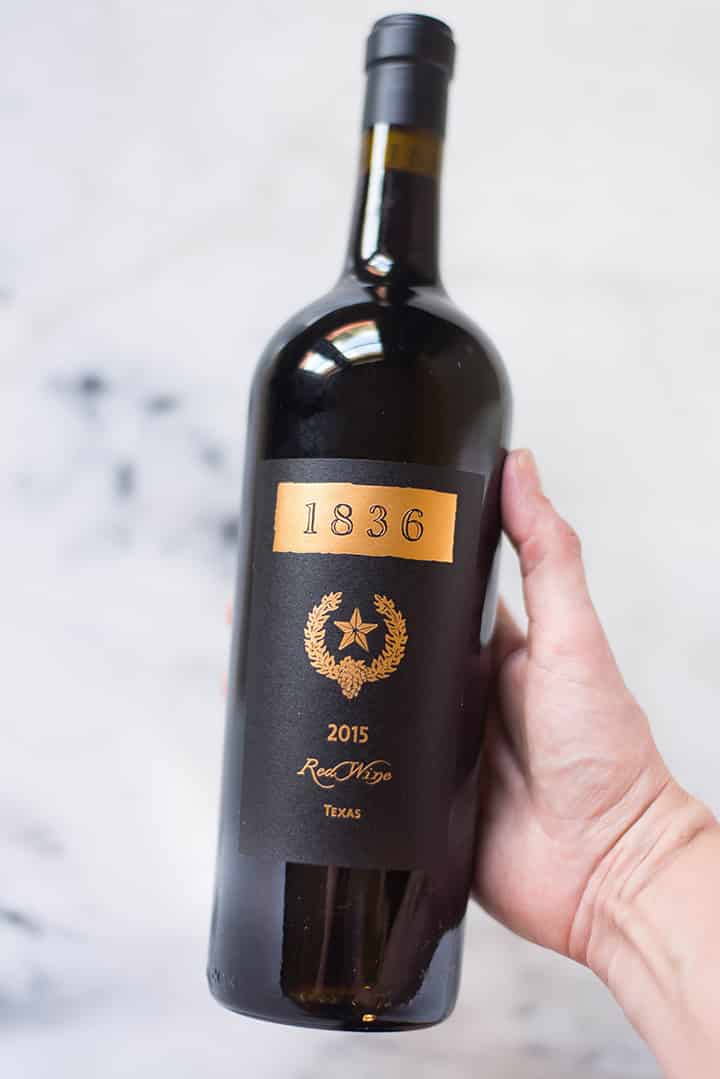 Hand holding an unopened bottle of Llano Estacado 1836 red wine, which is used in the Texas chili recipe.
