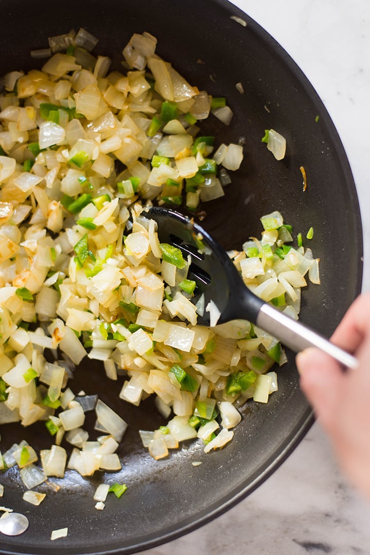 Close up image of a spoon stirring the onions and jalapenos on olive oil in a large skillet for the Spicy Quinoa and Black Bean Vegan Enchiladas filling.