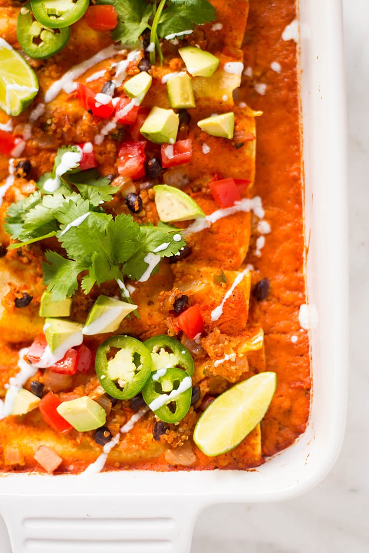 Close up image of the Spicy Quinoa and Black Bean Vegan Enchiladas: tortillas with the spicy quinoa and black bean enchilada filling and Homemade Vegan Enchilada Sauce topped with fresh cilantro, diced tomatoes and avocado, lime, and a little drizzle of non-dairy yogurt.