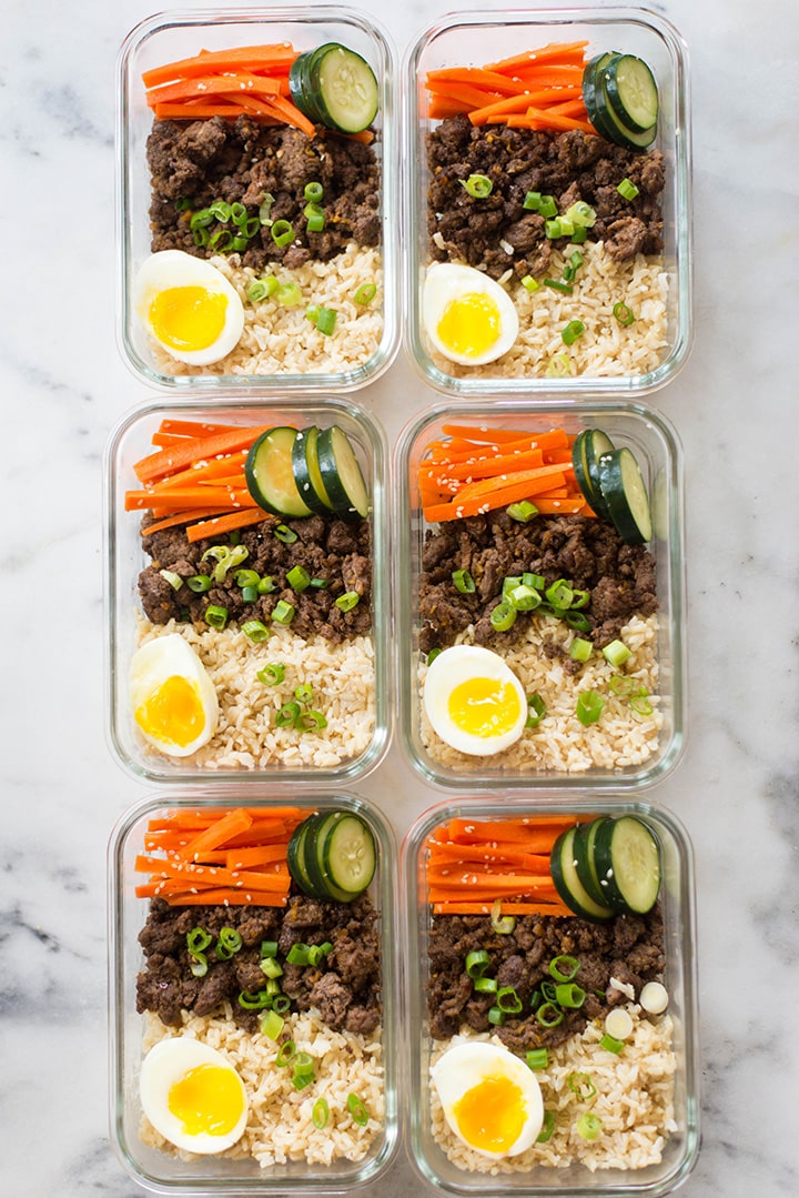 Overhead view of 6 meal prep containers containing meal prepped korean beef, carrot salad, steamed brown rice, and cucumbers plus a medium boiled egg.