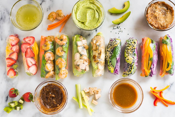 The 5 healthy spring roll recipes side by side in a line, including the rainbow spring roll, shrimp spring roll, garlic chicken spring roll, sesame avocado spring roll, and kiwi strawberry spring roll, next to their own dipping sauces and the different ingredients used in each one.