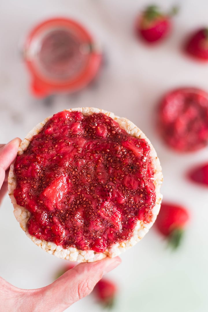 Hand holding a brown rice cake that has homemade strawberry jam spread over it and is ready to eat.
