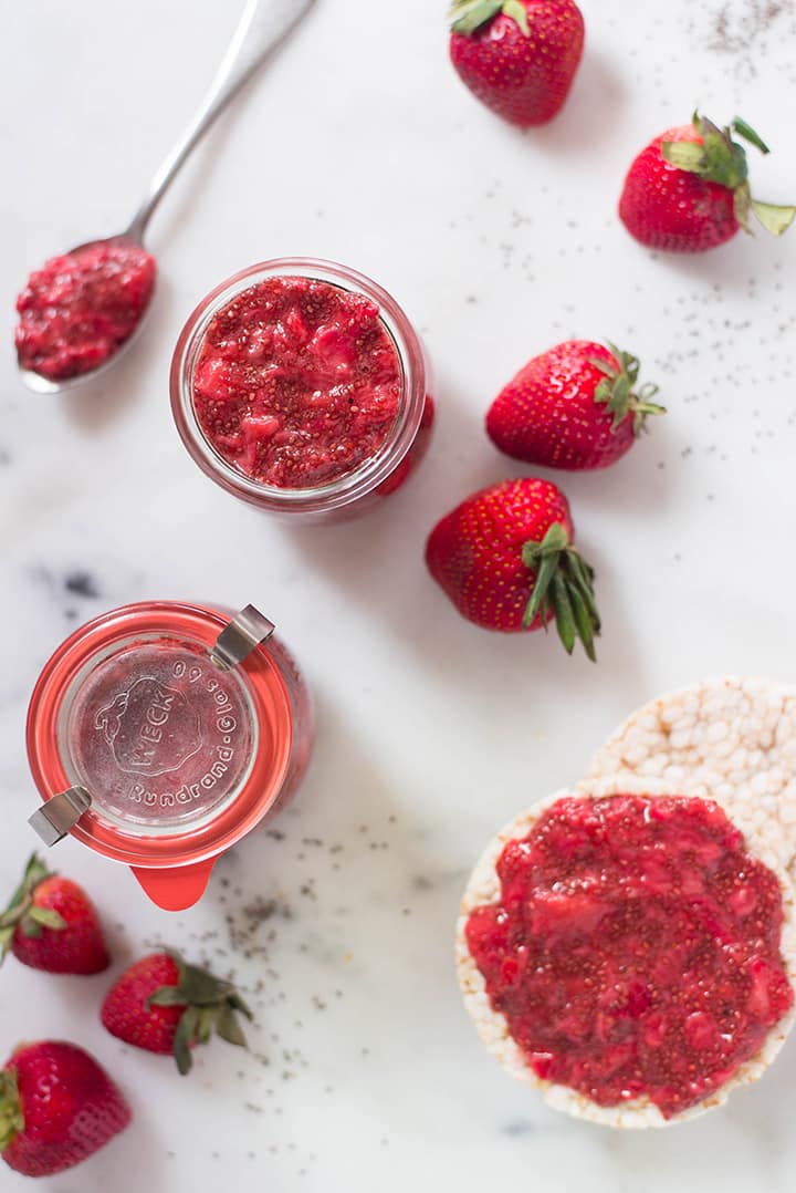 Overhead view of two jars of strawberry chia jam, siting next to fresh strawberries and a rice cake with strawberry chia jam spread.