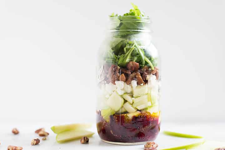Mason jar filled with ingredients for beet and goat cheese salad, setting next to pecans and sliced green apples.