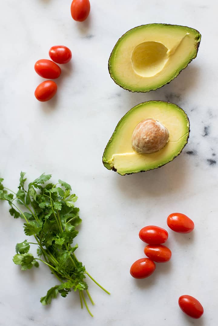 An overhead image of the ingredients for the Healthy Taco Salad with Creamy Cilantro Lime Dressing on a kitchen counter including an avocado, sliced in half, whole grape tomatoes and fresh cilantro.