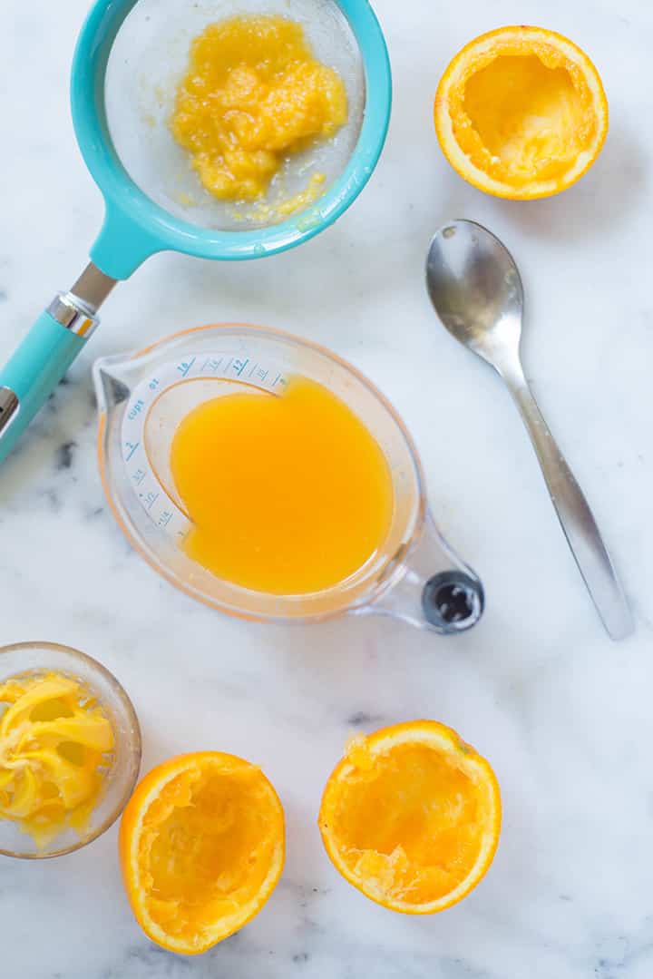 An overhead image of a kitchen counter with a mesh strainer and a glass pitcher with freshly squeezed and strained orange juice for the Caffeine-free Homemade Orange Soda.