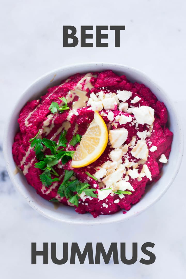 An overhead image of a bowl of Homemade Beet Hummus made from cooked chickpeas, beets, tahini, lemon juice, ground cumin, garlic, lemon zest and sea salt, garnished with crumbled feta, fresh parsley and a slice of lemon.