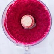 An overhead image of a food processor with Beet Hummus made from cooked beets, chickpeas, garlic, tahini, ground cumin, lemon zest and sea salt.