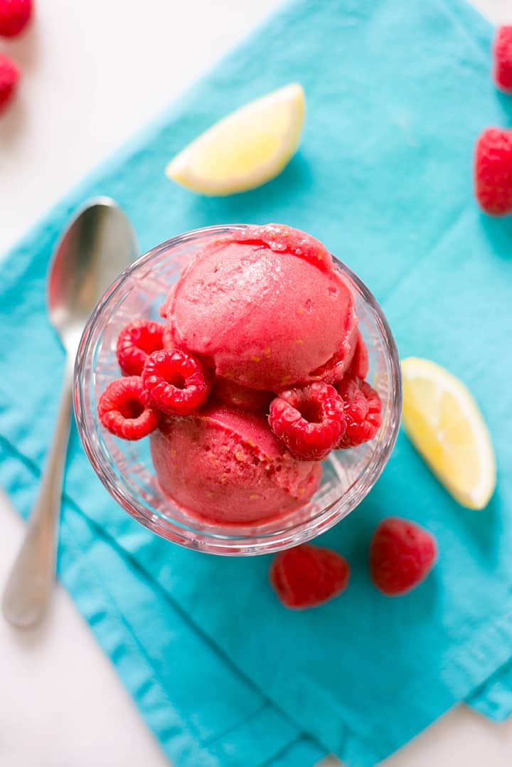 A close up of scoops of fresh Raspberry sorbet in a glass decorated with whole fresh raspberries.