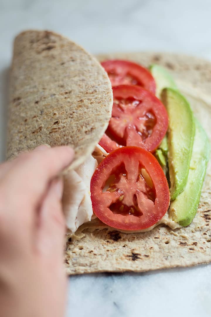 A close up image of making an Avocado Turkey Hummus Wrap, wrapping up a whole wheat tortilla filled with hummus, muenster cheese, smoked deli turkey breast, avocado, roma tomato and cucumber.