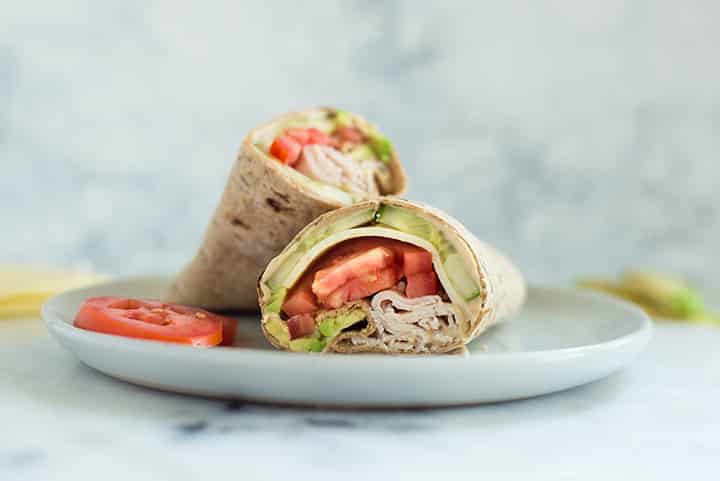 A close-up side image of an Avocado Turkey Hummus Wrap sliced in half, made with smoked deli turkey breast, hummus, avocado, muenster cheese, cucumber slices and roma tomatoes wrapped in a whole wheat wrap.