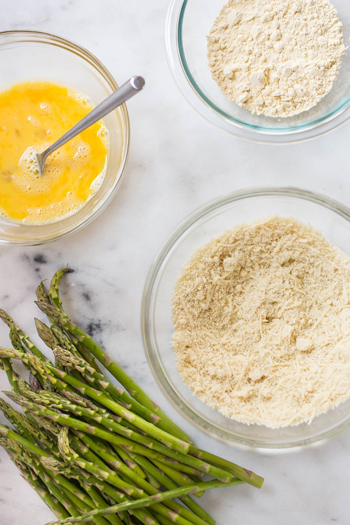 An overhead image of a kitchen counter with all the ingredients for Oven Baked Parmesan Asparagus Fries including fresh asparagus, eggs, chickpea flour, almond meal, grated parmesan, sea salt and pepper.
