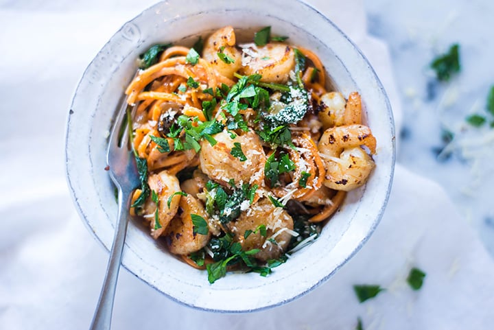 An overhead image of a serving of Creamy Garlic Tuscan Shrimp made with shrimp, garlic, yellow onion, low sodium chicken broth, sun dried tomato strips, unsweetened almond milk, baby spinach, parmesan, arrowroot starch and parsley served with sweet potato noodles.