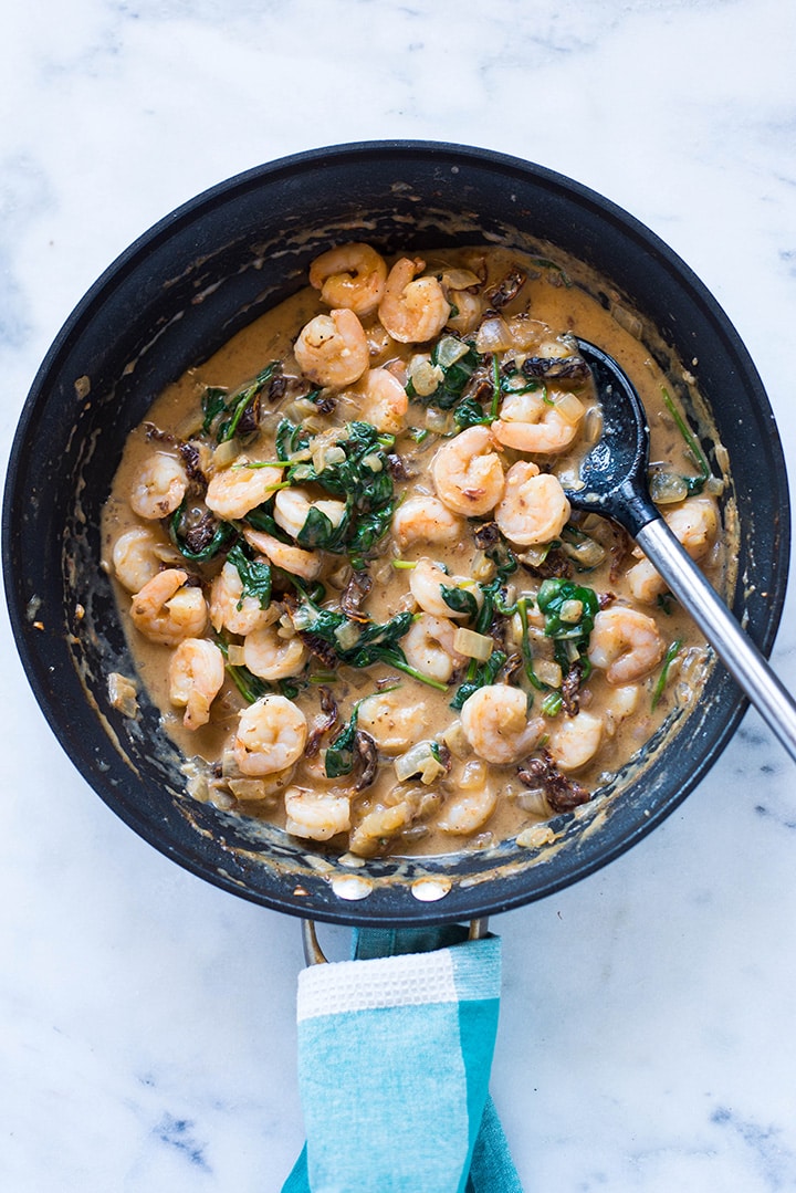 An overhead image of a large skillet with Creamy Tuscan Shrimp made with shrimp, garlic, yellow onion, low sodium chicken broth, sun dried tomato strips, unsweetened almond milk, baby spinach, parmesan and arrowroot starch.