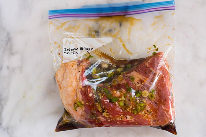 An overhead image of a sealable freezer bag with a tri tip steak in Sesame Ginger Tri Tip Marinade with sliced green onions, minced garlic, rice wine vinegar, raw honey, freshly grated ginger, crushed red pepper flakes, low sodium soy sauce, low sodium beef broth, olive oil and sesame oil.