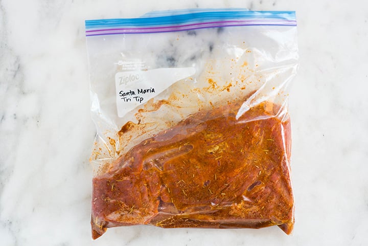 An overhead image of a sealable freezer bag with a tri tip steak in Santa Maria Tri Tip Marinade with sea salt, ground black pepper, garlic powder, paprika, onion powder, dried rosemary, cayenne pepper, dijon mustard, red wine vinegar and olive oil.