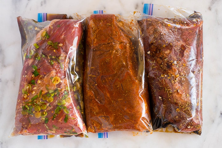An overhead image of three tri tip steaks in sealable freezer bags on a kitchen counter ready to be placed in the fridge to marinate, each in a different kind of tri tip marinade including Santa Maria Tri Tip Marinade, Balsamic Tri Tip Marinade and Sesame Ginger Tri Tip Marinade.