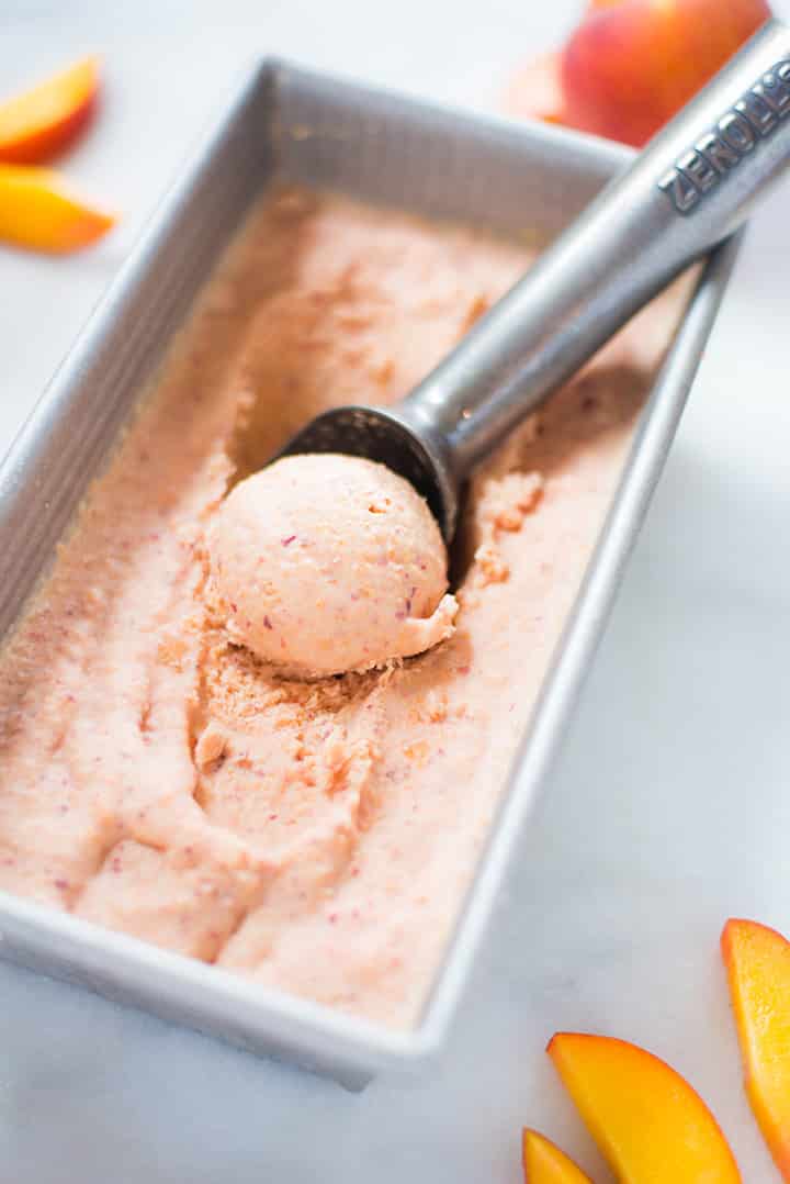 An overhead image of an ice cream scoop scooping up homemade Peach Ice Cream, made with ripe peaches, full-fat coconut milk, vanilla extract, maple syrup, lemon juice and cinnamon.