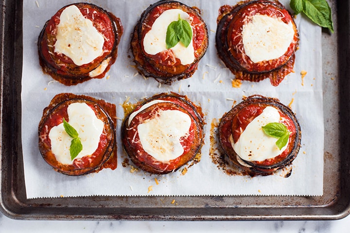 An overhead image of 6 stacks of Baked Eggplant Parmesan on a baking sheet, made with roasted eggplant, homemade tomato sauce, mozzarella and grated parmesan.