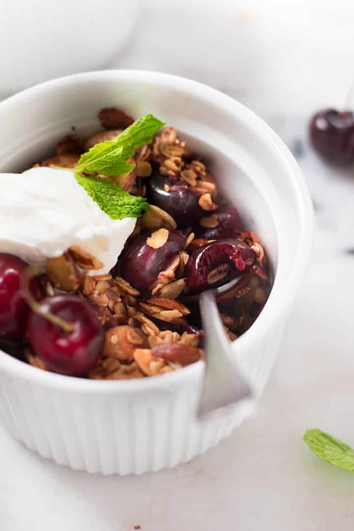 A side close up image of a ramekin with baked Fresh and Healthy Cherry Crisp, served with a dollop of plain Greek yogurt and decorated with fresh mint.