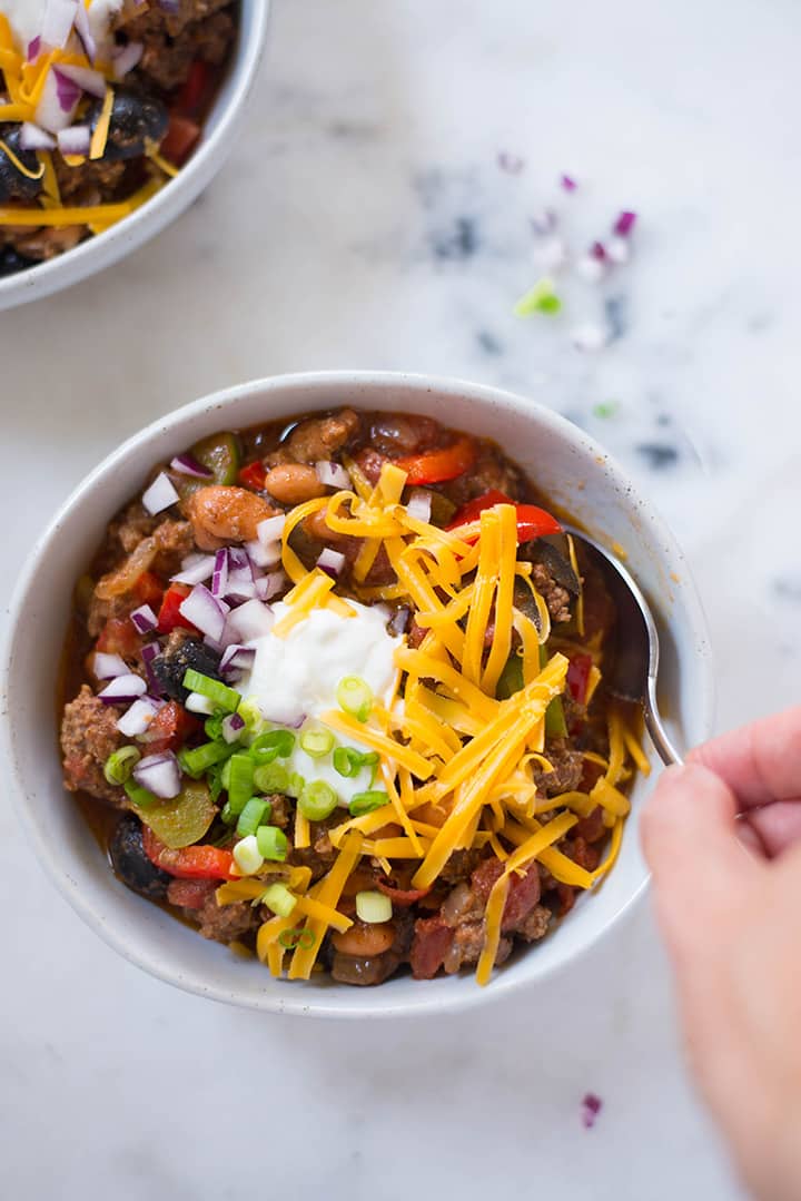 An overhead image of a bowl of Homemade Chili on a kitchen counter, made with ground beef, pinto beans, green and red bell pepper, canned diced tomatoes, black olives, yellow onion, garlic, olive oil and tomato sauce, garnished with grated cheddar cheese, green onions and greek yogurt.