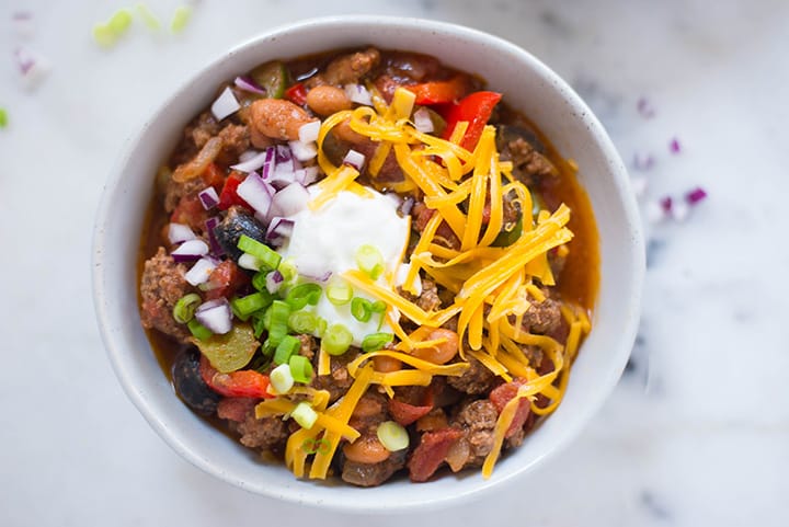 An overhead image of a bowl of Homemade Chili made with ground beef, pinto beans, green and red bell peppers, canned diced tomatoes, black olives, yellow onion, garlic, olive oil and tomato sauce, garnished with grated cheddar cheese, green onions and greek yogurt.