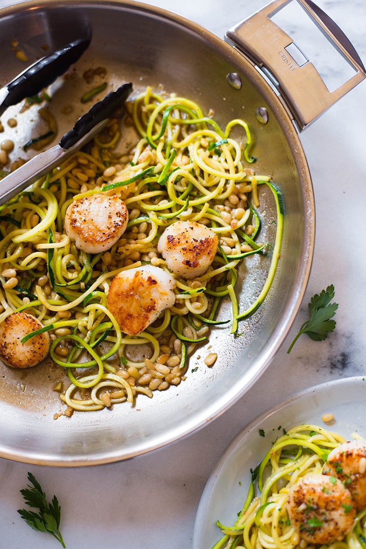 Pan Seared Scallops with zucchini noodles and roasted pine nuts in the Cristel Frying pan ready to be plated. Near the pan you can see an already plated serving of Pan Seared Scallops.