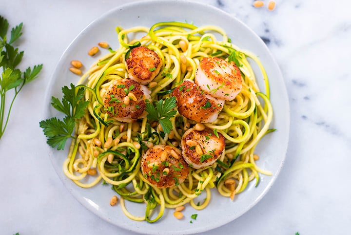 A serving of Pan Seared Scallops over zucchini noodles topped with roasted pine nuts.