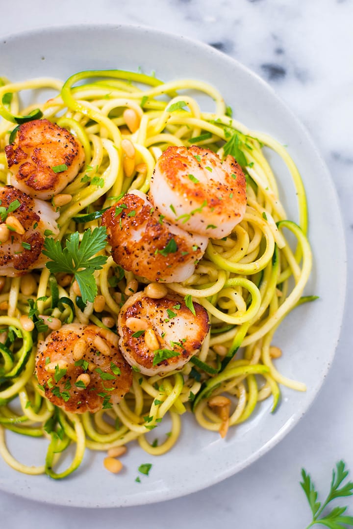 Close up of Pan Seared Scallops on a bed of zucchini noodles topped with pine nuts and parsley on a plate ready to be eaten.