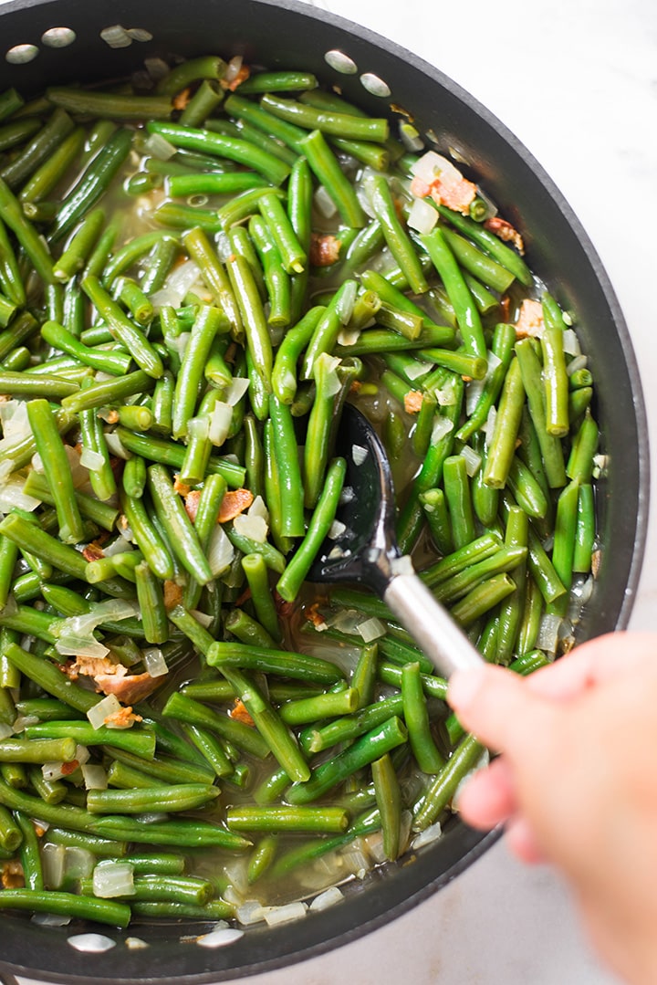 An overhead image of a spoon stirring the Southern Green Beans in a pan, made with fresh green beans, cooked bacon, low sodium chicken broth, onion and garlic.