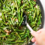 A close up image of a spoon stirring Southern Green Beans made with green beans, cooked bacon, low sodium chicken broth, onion and garlic.