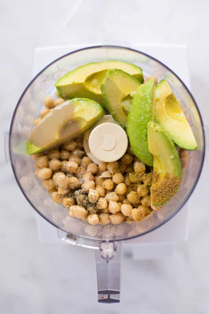 Separated ingredients including avocados, chickpeas, tahini, olive oil, sea salt, pepper in the food processor ready to be mixed and transformed into Healthy Avocado Hummus.