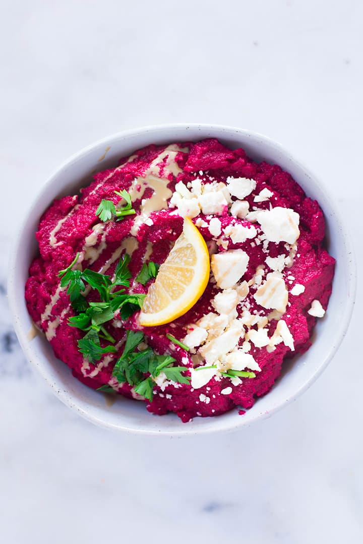 Overhead view of a white bowl containing beet hummus garnished with feta cheese, parsley, and a lemon slice ready to be served.