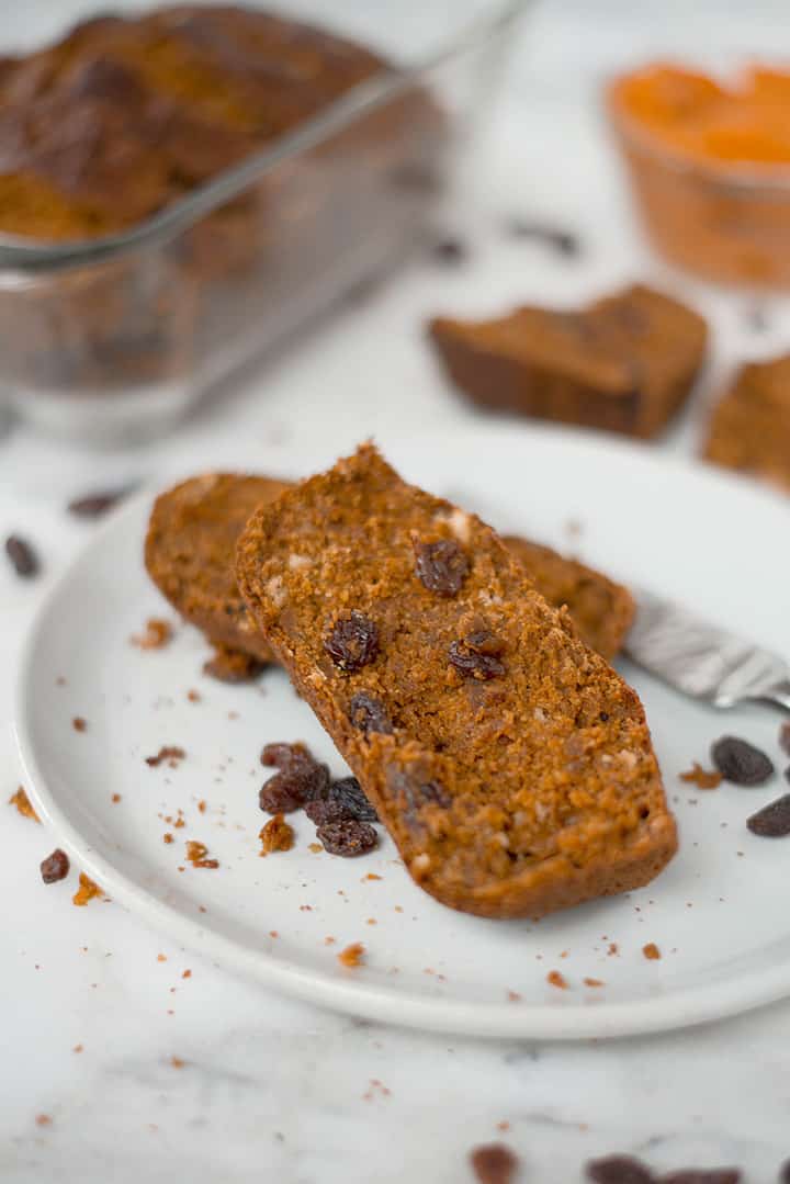 2 slices of Healthy Pumpkin Bread on a serving plate. In the background can be seen other slices of pumpkin bread, the loaf pan with the remaining pumpkin bread, and a mixing bowl with pumpkin puree.