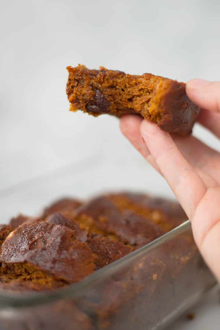 Close up of a hand holding a slice of Healthy Pumpkin Bread. In the background can be seen the rest of the pumpkin bread that is still in the loaf pan.