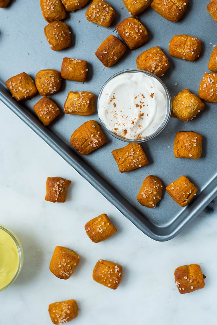 Overhead image of Soft Pretzel Bites with Sweet Potato Dough on a baking tray with the sweet yogurt-based dipping sauce placed in between them. Some pretzel bites are place on the table.