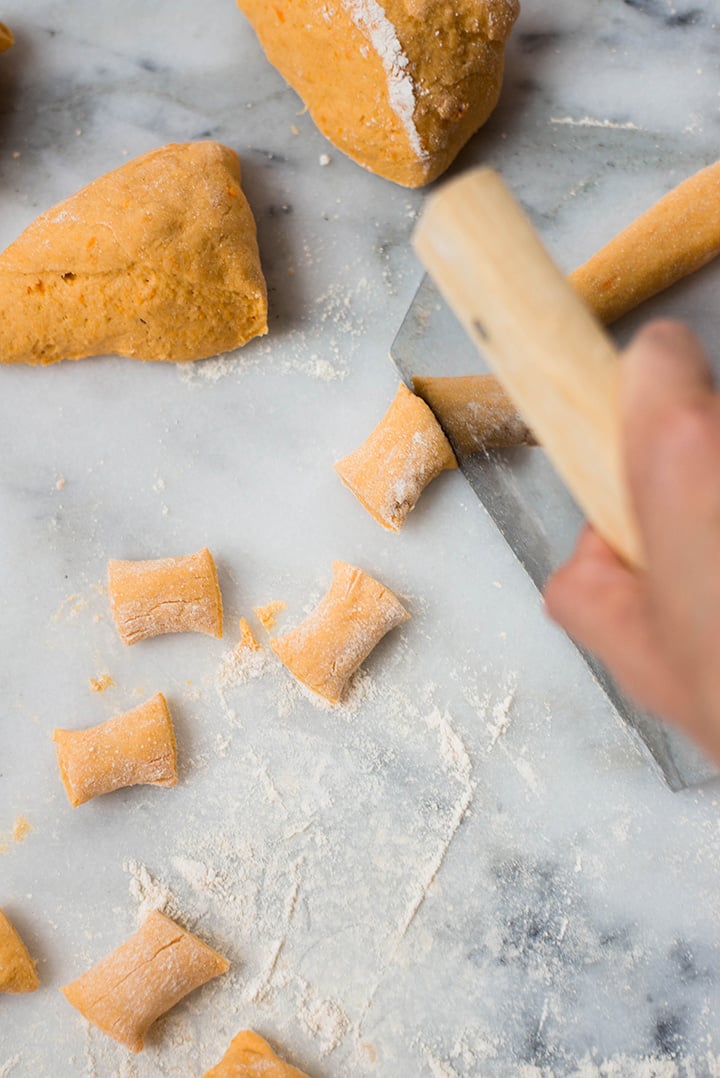 A skinny rope of sweet potato dough being cut into pretzel bites. In the background can be seen 2 pieces of sweet potato dough that haven't been rolled into skinny rope shape.