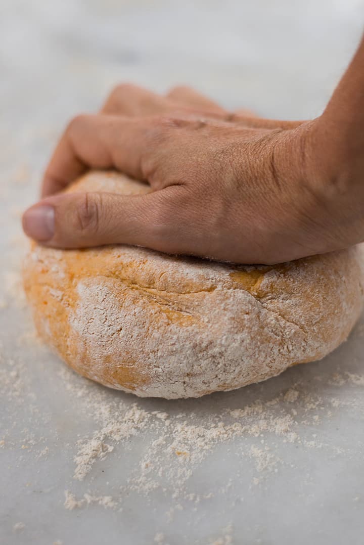 Close up of a hand kneading sweet potato dough on a floured surface.