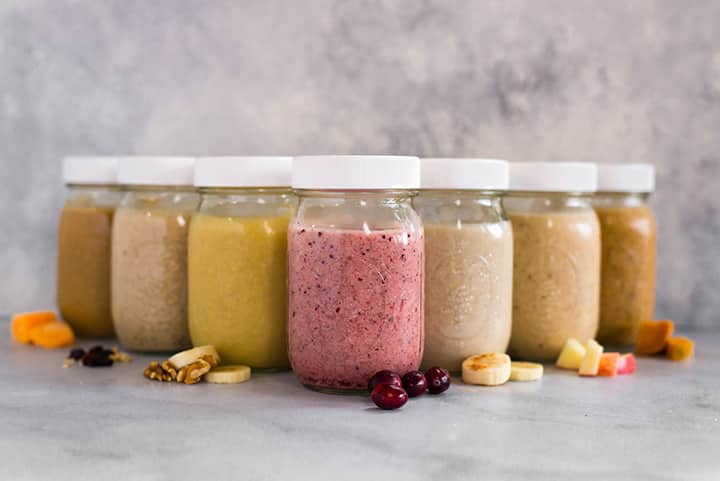 Side view of 7 mason jars with Easy Make Ahead Smoothies for Fall lined up, with fruit and veggies beside the jars, including pumpkin, berries, and bananas.