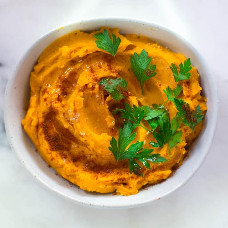 Overhead view of a white bowl full of Healthy Sweet Potato Hummus, topped with parsley and olive oil.