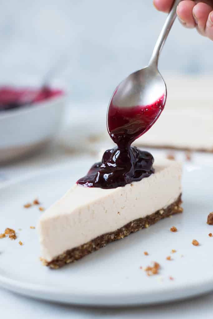 Hand pouring cherry topping with a spoon over a slice of vegan no bake cheesecake.