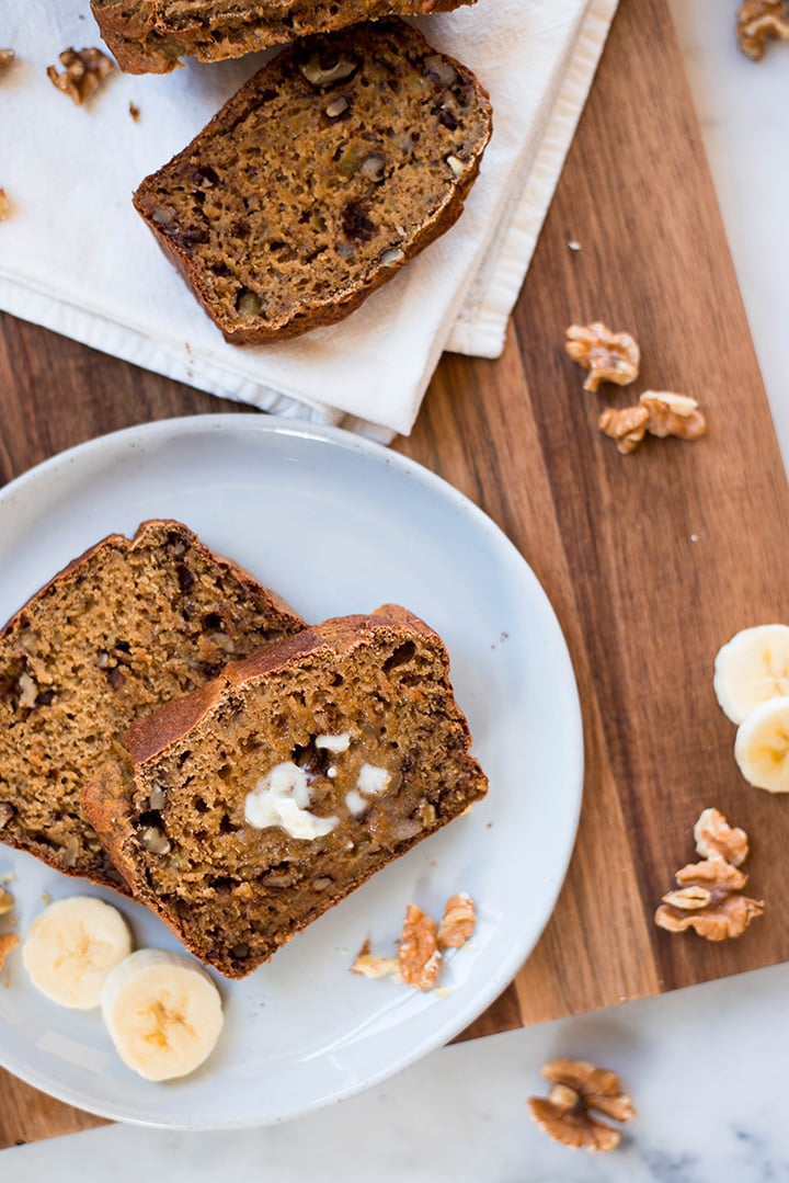2 serving plates with 2 slices of banana bread on each plate. Slices of bananas and walnuts are placed on and near the plates for garnish and as decoration.