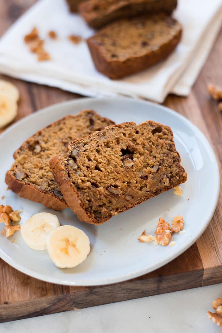 Close up of a serving plate with 2 slices of banana bread garnished with slices of fresh bananas and walnuts.