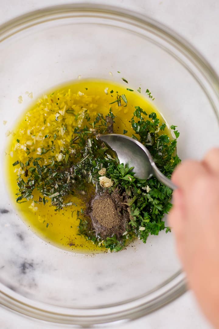 Close up of a hand mixing the ingredients for the chicken marinade including olive oil, lemon juice, apple cider vinegar, parsley, rosemary, garlic, salt, and pepper.