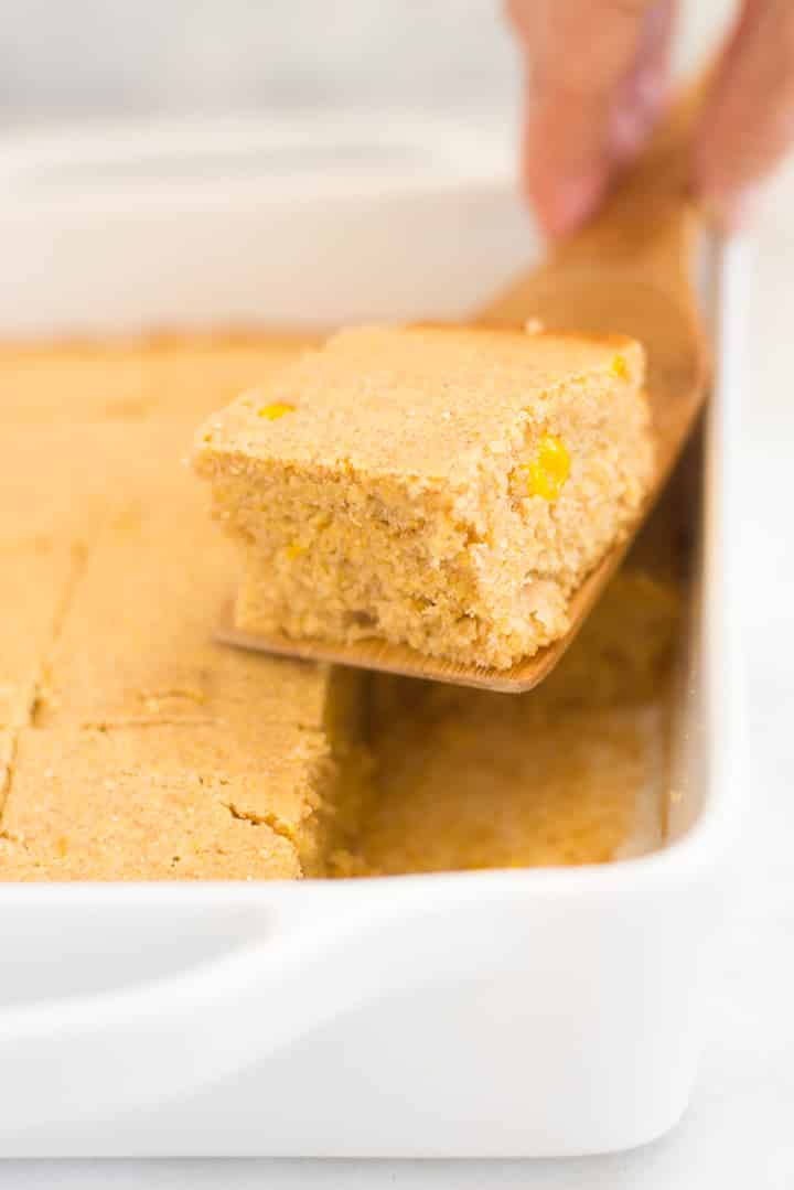 Close up side view of a white rectangular dish which contains sweet cornbread. A wooden spatula is being used to remove a piece of the bread.