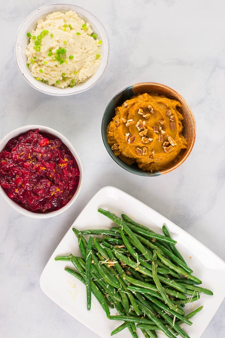 Overhead image of 3 bowls: one bowl contains cranberry sauce, one contains sweet potatoes, one mashed potatoes, and there is a platter of green beans.