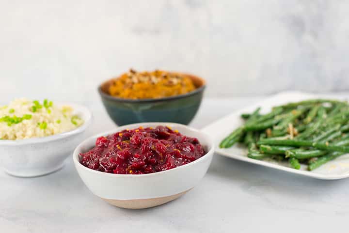 Side view of 4 Healthy Thanksgiving Side Dishes. The cranberry sauce, mashed sweet potatoes, and cauliflower mashed potatoes are in serving bowls while the garlic parmesan garlic beans are placed on a serving plate.
