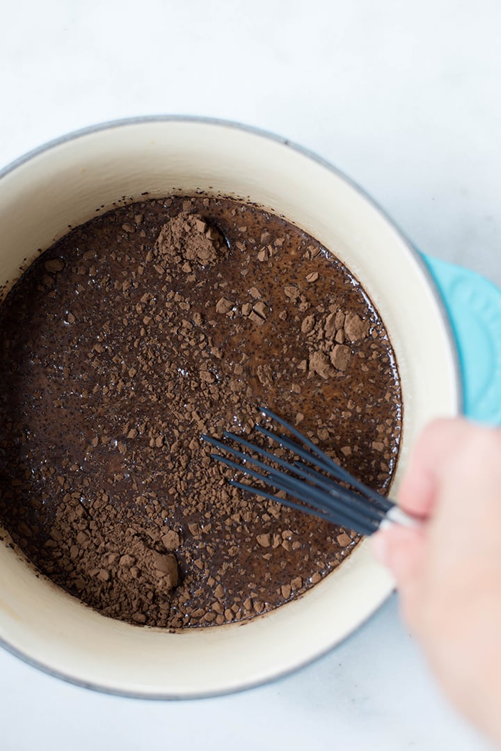Mixing the ingredients for a mocha latte including strongly-brewed coffee, cocoa powder, almond milk, coconut milk, and vanilla extract in a saucepan with a whisk.