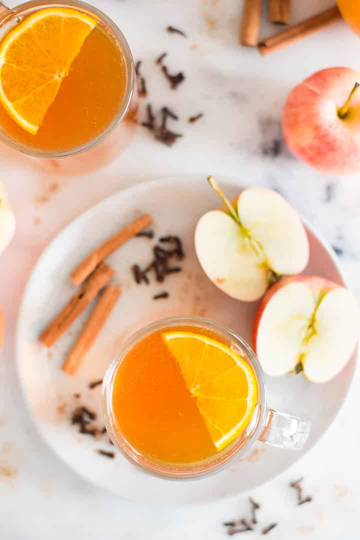 View from the top of 2 apple cider mugs. One of the mugs is on a serving plate. Both apple cider mugs are garnished with orange slices and in the background can be seen some of the ingredients from the apple cider recipe.